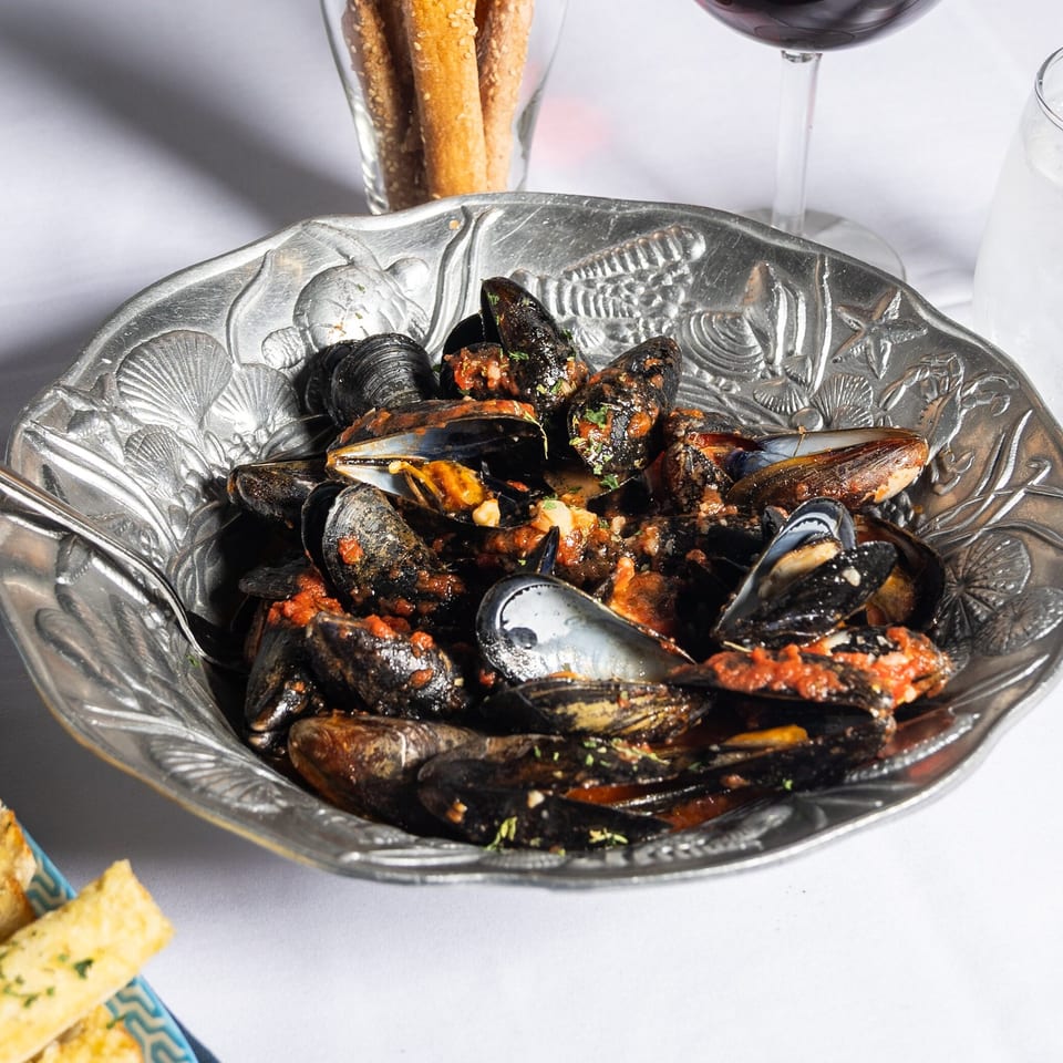 Steamed mussels in a rich red sauce, served in an ornate silver bowl with breadsticks and wine.