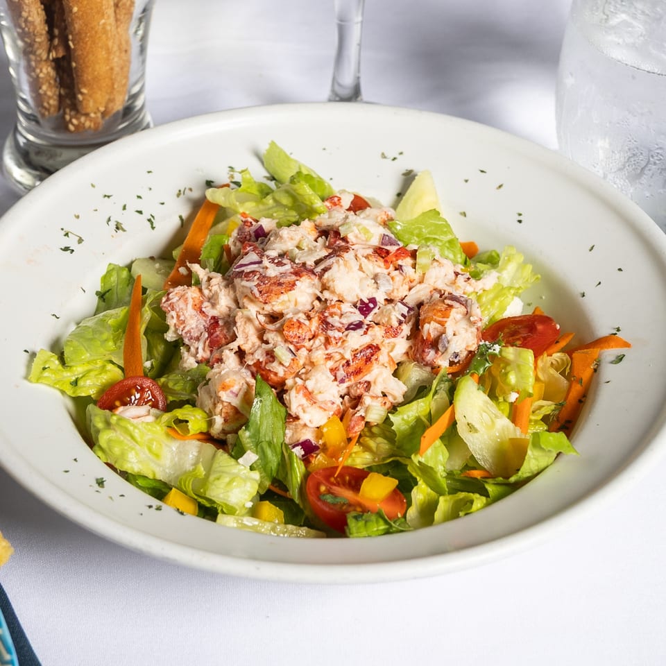 A vibrant lobster salad with fresh greens, carrots, tomatoes, and red onion in a white bowl.