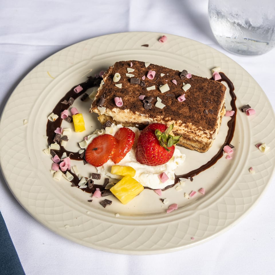 Tiramisu dessert with chocolate drizzle, whipped cream, strawberries, pineapple, and chocolate shavings.