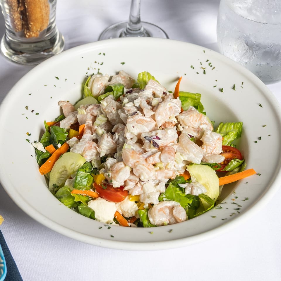 Shrimp salad with lettuce, tomatoes, carrots, and cucumbers in a white bowl on a white tablecloth.