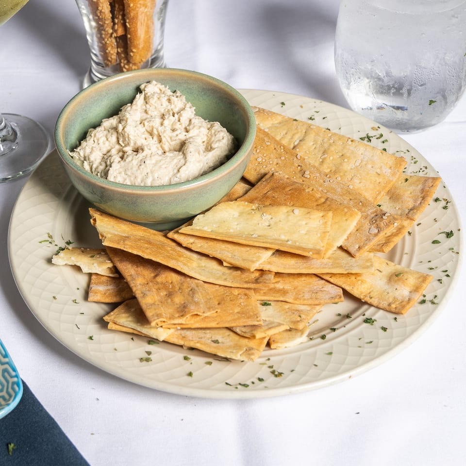 A bowl of creamy dip with flatbread crackers on a plate, alongside drinks on a white tablecloth.