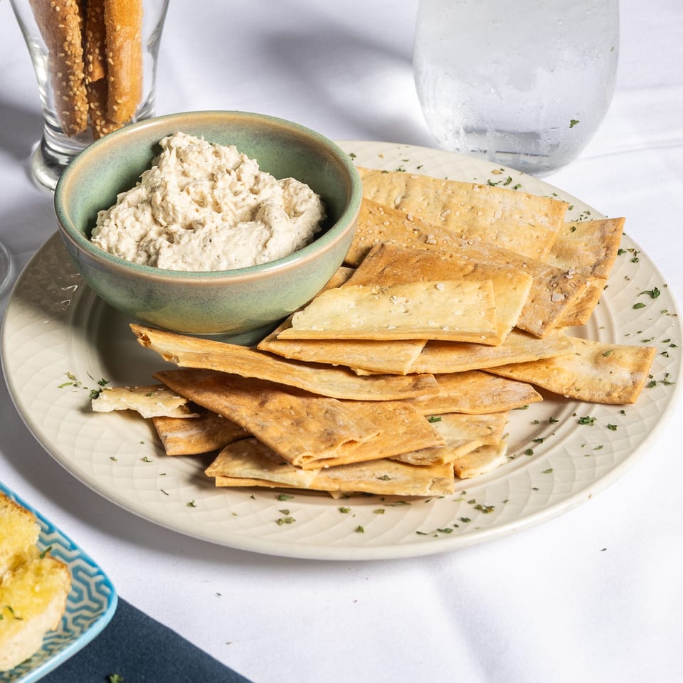 A bowl of white dip with flatbread crackers on a plate, alongside breadsticks and drinks.