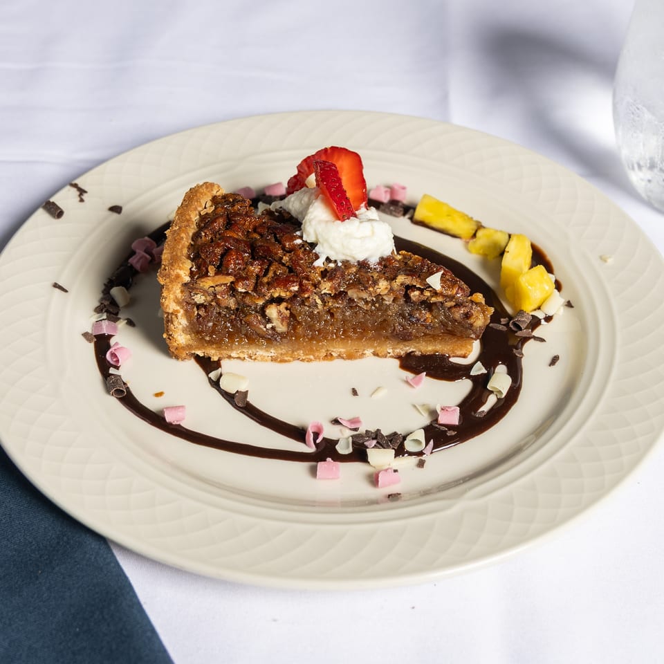 A slice of pecan pie with whipped cream, strawberry, pineapple, and chocolate drizzle on a white plate.