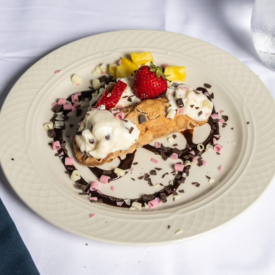 A cannoli dessert on a white plate, drizzled with chocolate, topped with strawberries, pineapple, and chocolate shavings.