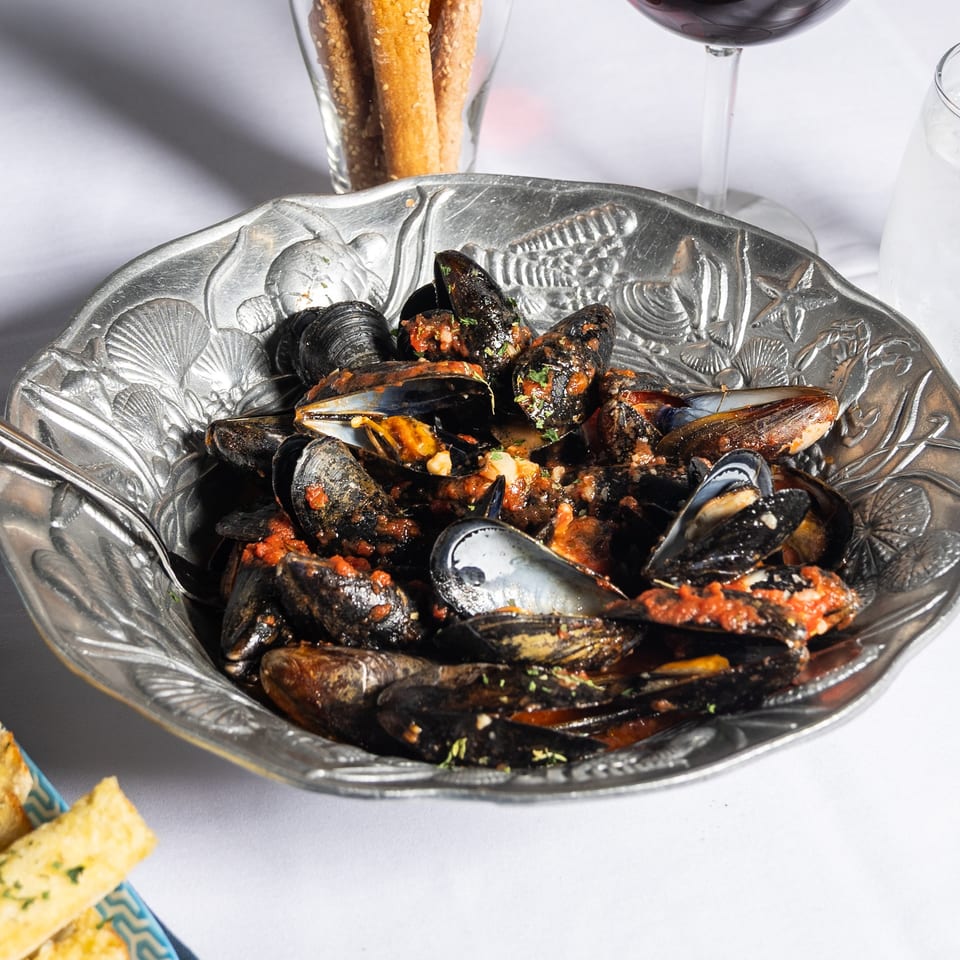 Steamed mussels in a rich red sauce, served in an ornate silver bowl with breadsticks and wine.
