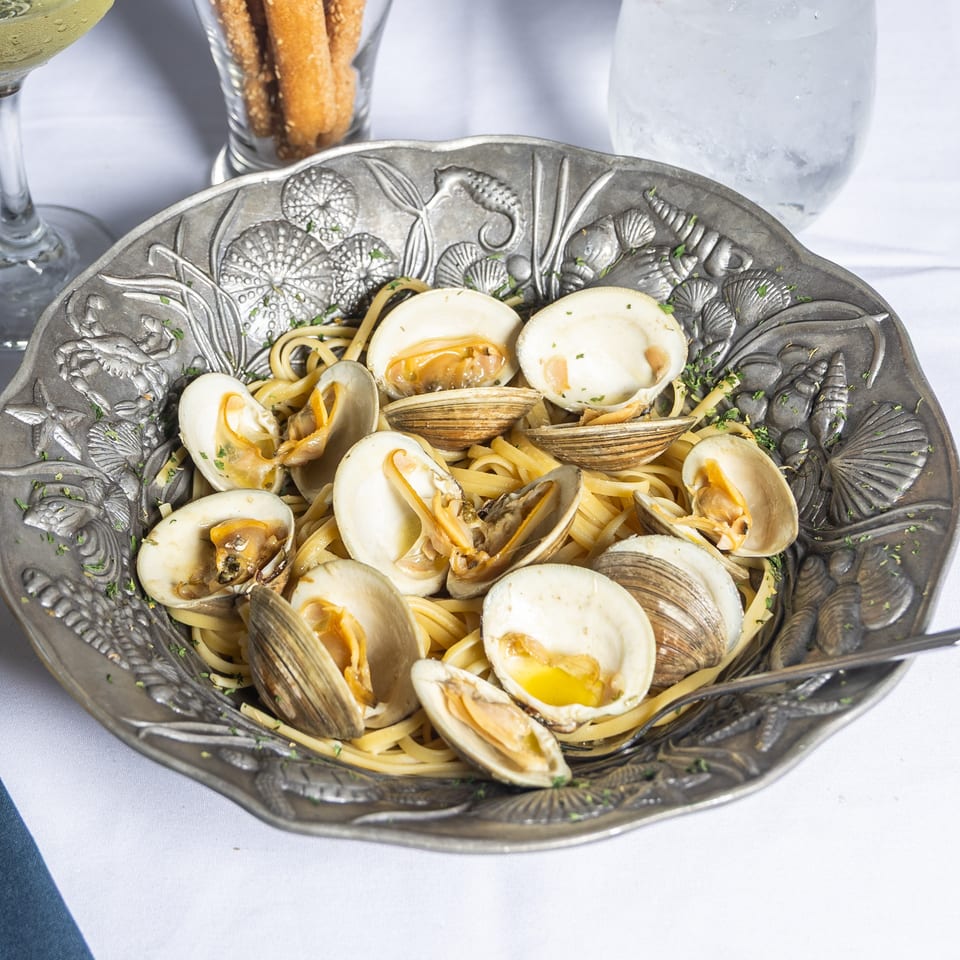 Linguine with clams in a decorative metal bowl, with wine and breadsticks on a white tablecloth.