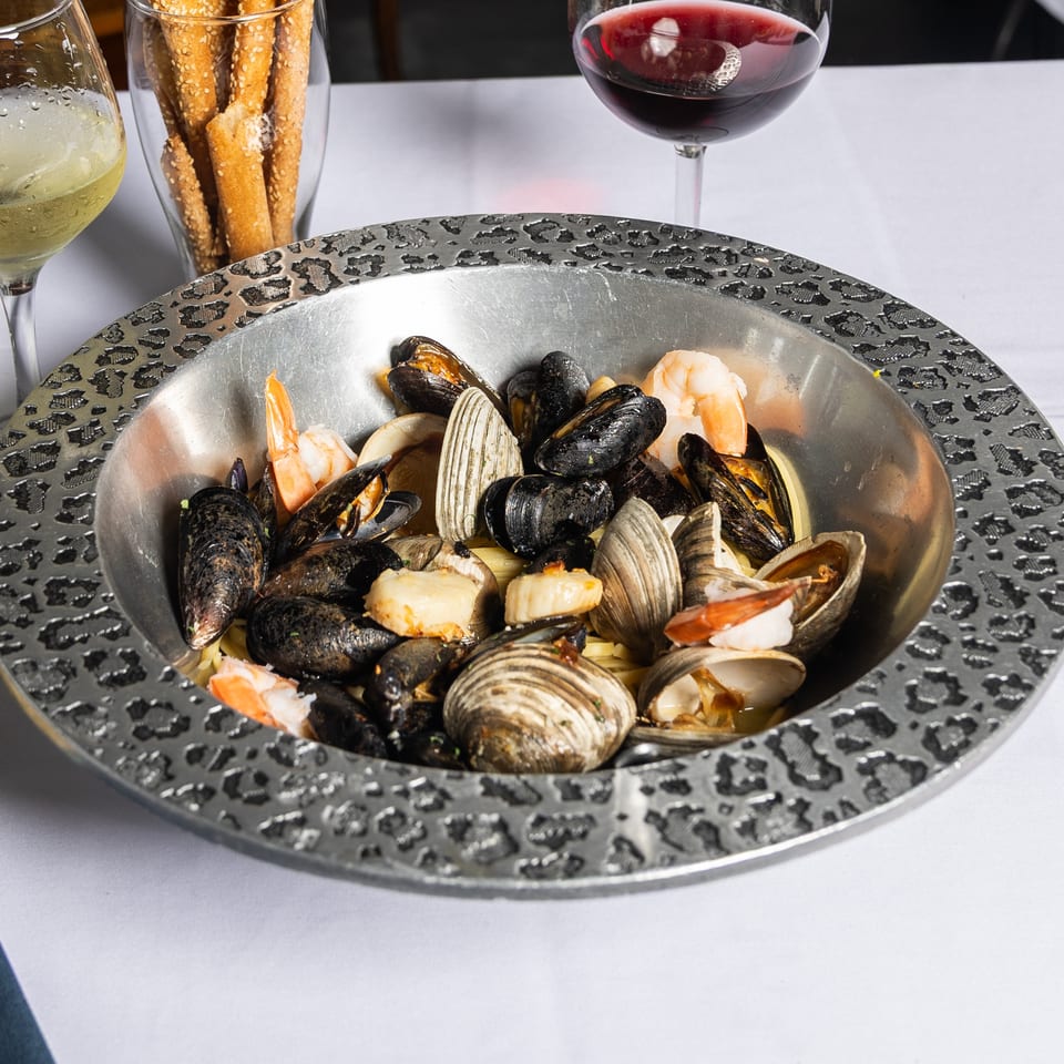 Seafood pasta with mussels, clams, and shrimp in a decorative metal bowl, with wine and breadsticks.