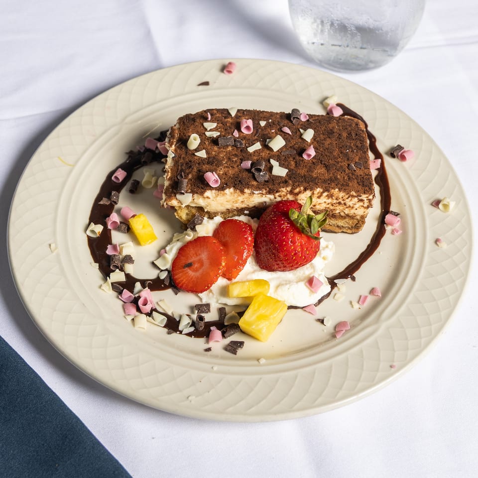 Tiramisu dessert with chocolate shavings, strawberries, pineapple, and whipped cream on a white plate.