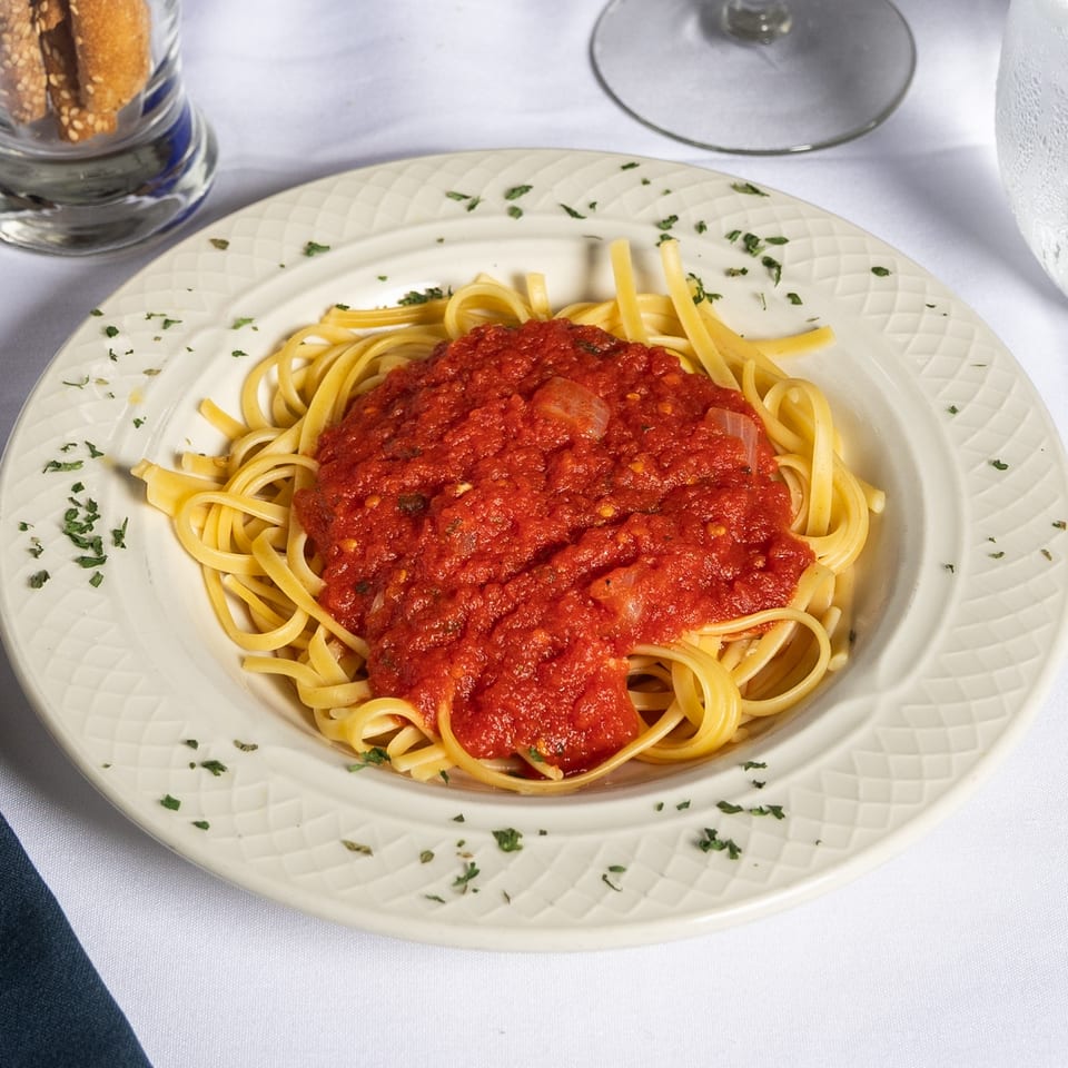 Linguine pasta with red tomato sauce and parsley on a white plate, with breadsticks and water glass.